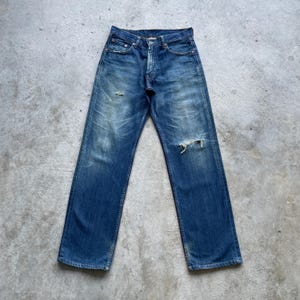 May include: Faded blue denim jeans with distressed details, including a ripped knee. The jeans have a classic five-pocket design and a button and zipper closure. The jeans are laid flat on a concrete surface.