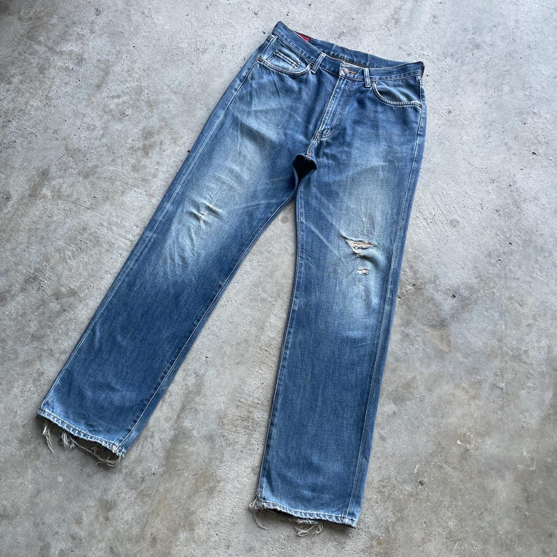 Pu&ograve; includere: Jeans in denim blu sbiadito con un aspetto invecchiato. I jeans hanno un classico design a cinque tasche, una chiusura con bottoni e un aspetto usurato con alcuni strappi. L'orlo &egrave; sfilacciato, aggiungendo all'estetica vintage.