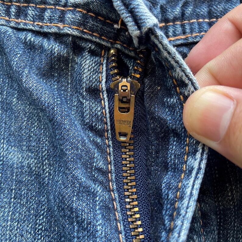 Puede incluir: Primer plano de una cremallera dorada de unos vaqueros azules. La cremallera tiene grabado el nombre de la marca "EDWIN". Los vaqueros tienen costuras visibles y una textura vaquera cl&aacute;sica.