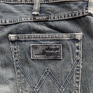 May include: Close-up of the back pocket of blue denim jeans. The Wrangler brand is visible on a small tag above the pocket and on the leather patch. White stitching outlines the pocket and forms the iconic 'W' design.