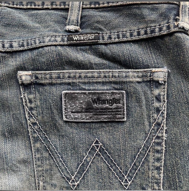May include: Close-up of the back pocket of blue denim jeans. The Wrangler brand is visible on a small tag above the pocket and on the leather patch. White stitching outlines the pocket and forms the iconic 'W' design.