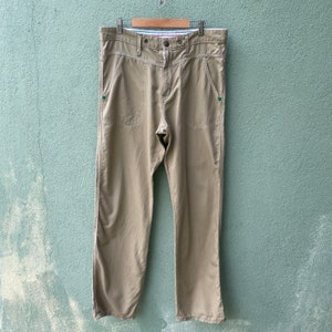 May include: Khaki-coloured trousers with a button closure and pockets. The trousers are hanging on a wooden hanger. The trousers have a straight leg design and are made of a lightweight material.