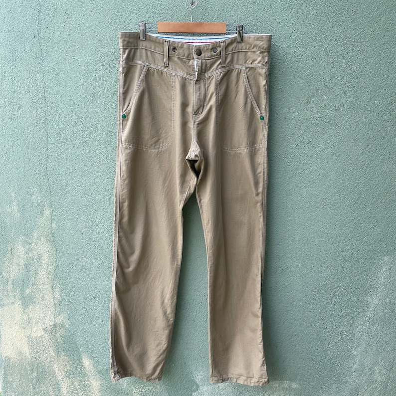 May include: Khaki-coloured trousers with a button closure and pockets. The trousers are hanging on a wooden hanger. The trousers have a straight leg design and are made of a lightweight material.