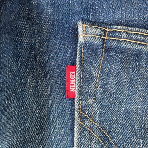 Puede incluir: Primer plano de vaqueros azules con un bolsillo trasero y una etiqueta roja de Edwin. El bolsillo est&aacute; delineado con costuras color canela. El denim tiene una apariencia texturizada.