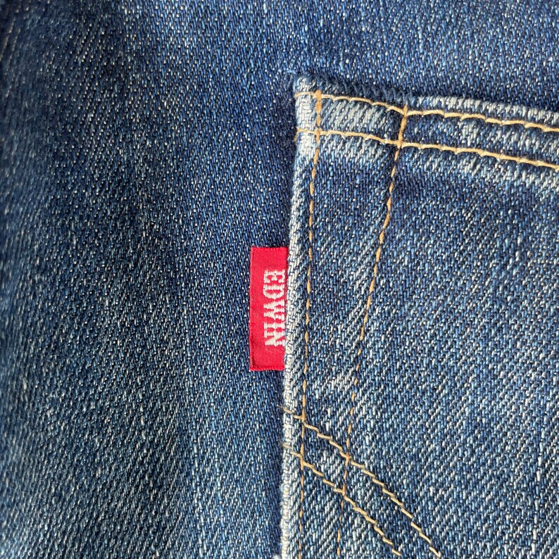 Puede incluir: Primer plano de vaqueros azules con un bolsillo trasero y una etiqueta roja de Edwin. El bolsillo est&aacute; delineado con costuras color canela. El denim tiene una apariencia texturizada.