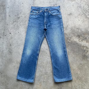 May include: Blue denim jeans with a classic five-pocket design. The jeans have a faded wash and a straight leg fit. The jeans are button-fly and have a worn appearance. The jeans are a vintage style.