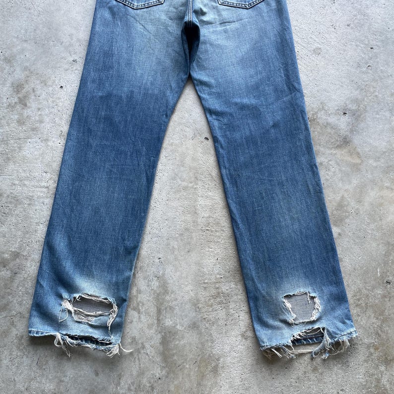 Pu&ograve; includere: Jeans di denim blu sbiadito con dettagli strappati. I jeans hanno un taglio dritto e presentano grandi fori strappati vicino alle caviglie. Il denim mostra segni di usura e sbiadimento, conferendo loro un aspetto vintage.