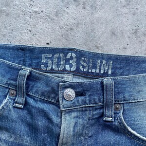 Puede incluir: Primer plano de unos vaqueros azules con el texto "503 SLIM" en la cintura. Los vaqueros tienen cierre de bot&oacute;n y trabillas para el cintur&oacute;n. El tejido vaquero tiene un aspecto descolorido y desgastado.