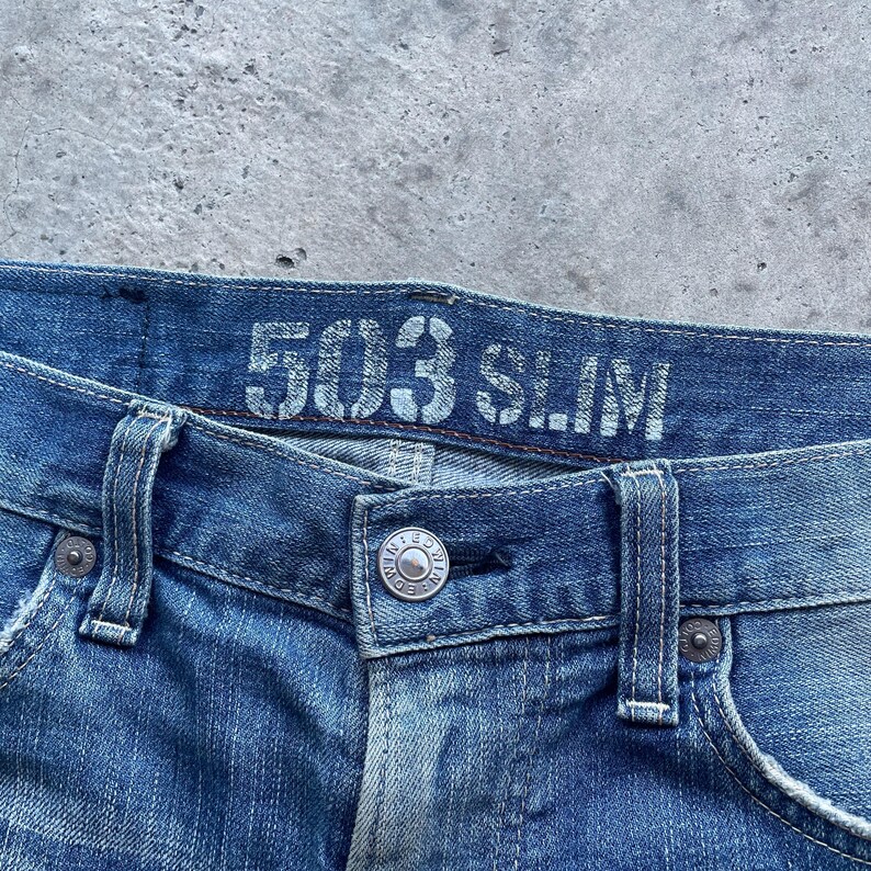 Puede incluir: Primer plano de unos vaqueros azules con el texto "503 SLIM" en la cintura. Los vaqueros tienen cierre de bot&oacute;n y trabillas para el cintur&oacute;n. El tejido vaquero tiene un aspecto descolorido y desgastado.
