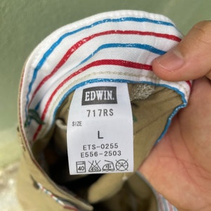 May include: A close-up of an Edwin clothing label, showing the brand name, style number, and size L. The label is white with black text. The inside of the garment has a striped pattern in red, white, and blue.