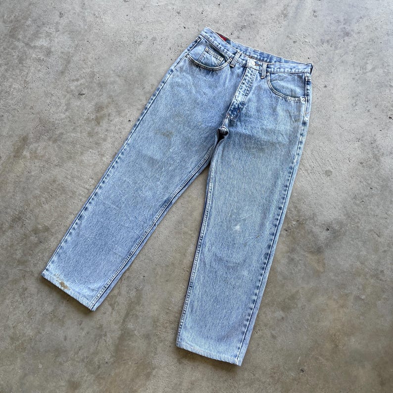 May include: Light blue acid wash denim jeans. The jeans have a classic five-pocket design with a button and zipper closure. The denim has a faded, worn appearance, with some light discoloration. The jeans are laid flat on a concrete surface.