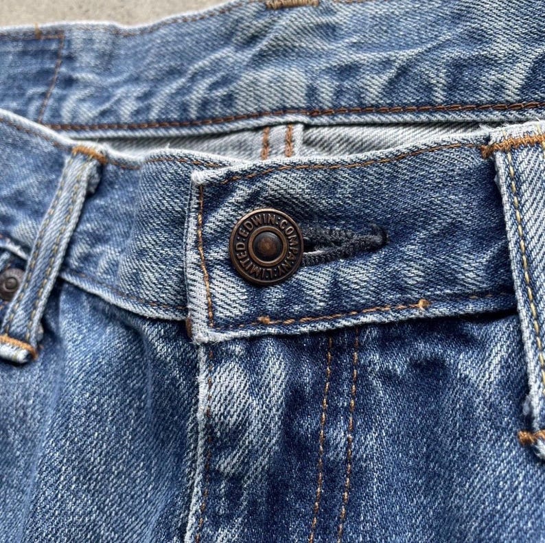 May include: Close-up of blue denim jeans, focusing on the button closure. The bronze-coloured button is embossed with "EDWIN CO. LIMITED". The denim fabric shows wear and tear, with visible stitching and a classic blue hue.