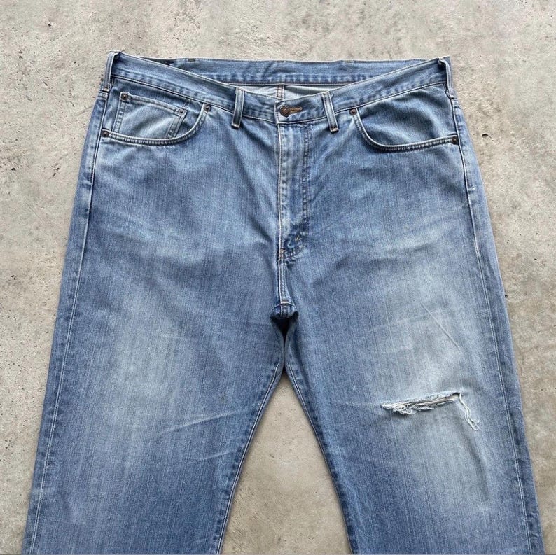 K&ouml;nnte beinhalten: Hellblaue Jeans mit einem aufgerissenen Knie. Die Jeans haben ein klassisches F&uuml;nf-Taschen-Design, einen Knopf- und Rei&szlig;verschluss sowie G&uuml;rtelschlaufen. Der Denim hat ein verwaschenes Aussehen.