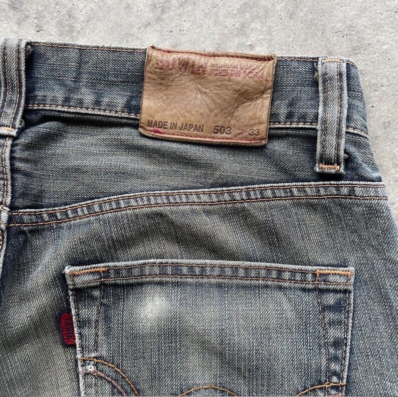 Puede incluir: Primer plano de vaqueros de mezclilla azul descoloridos. El bolsillo trasero tiene una etiqueta roja. Un parche de cuero marr&oacute;n en la cintura dice "EDWIN" y "MADE IN JAPAN 503 33". Los vaqueros tienen costuras visibles y un aspecto desgastado.