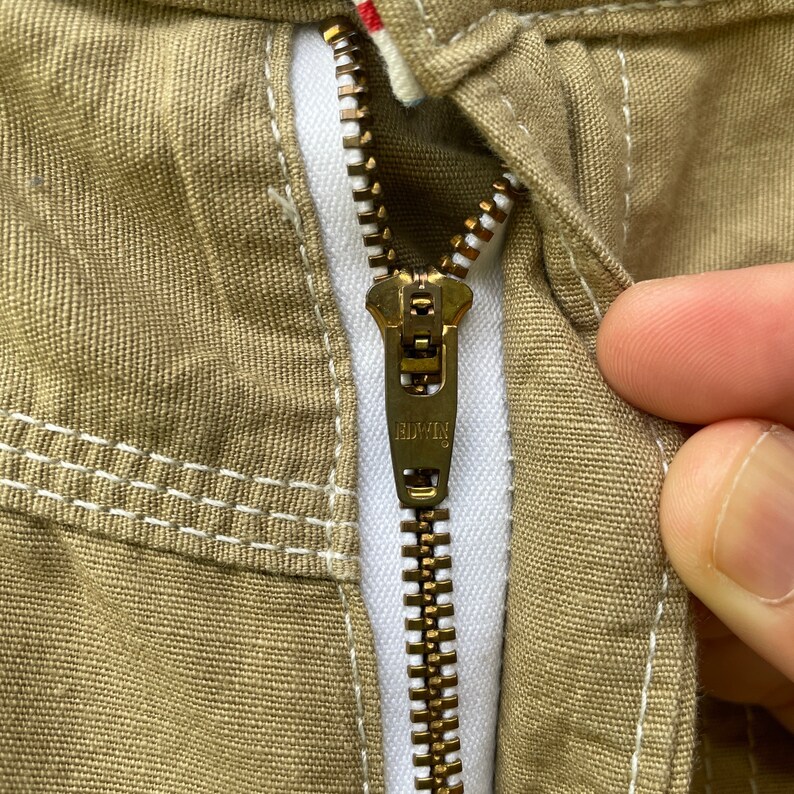 May include: Close-up of a khaki-coloured garment zipper. The zipper is gold-toned with the brand name "EDWIN" visible on the pull tab. The fabric has white stitching and a red and white detail near the top.