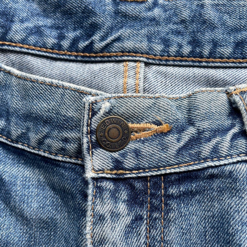 May include: Close-up of blue denim jeans, showing the button and buttonhole. The button is a dark metal colour with the text "EDWIN CO." around the edge. The stitching is a light brown colour.