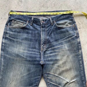 K&ouml;nnte beinhalten: Blaue Jeans aus Denim mit einem gelben Ma&szlig;band &uuml;ber der Taille. Die Jeans weisen Gebrauchsspuren auf, mit verblichenen Stellen und einem Riss am rechten Bein. Die Taille misst etwa 46 cm.