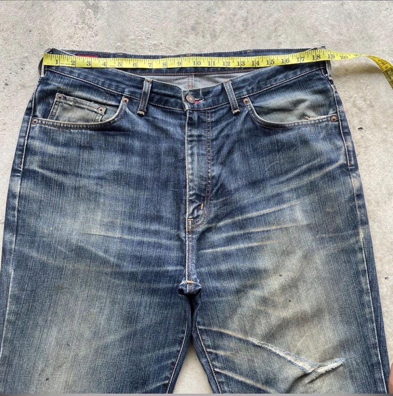 K&ouml;nnte beinhalten: Blaue Jeans aus Denim mit einem gelben Ma&szlig;band &uuml;ber der Taille. Die Jeans weisen Gebrauchsspuren auf, mit verblichenen Stellen und einem Riss am rechten Bein. Die Taille misst etwa 46 cm.