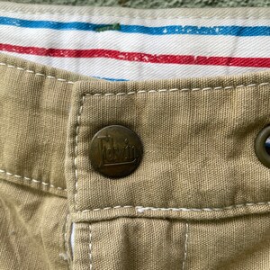 May include: Close-up of khaki fabric with a metal button bearing the brand name "Edwin." White stitching is visible around the button. The interior lining features red and blue stripes.
