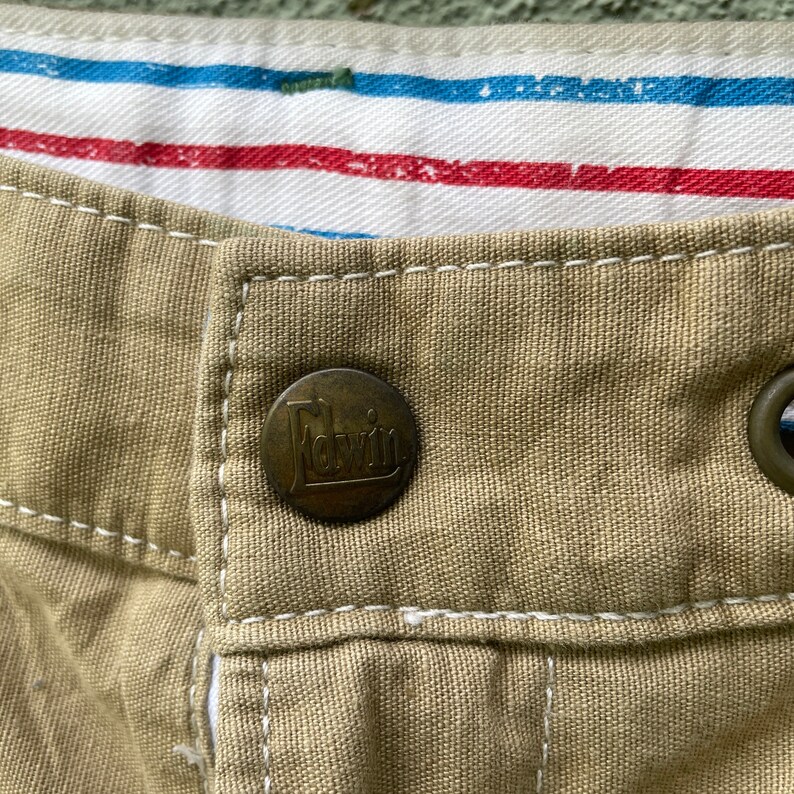 May include: Close-up of khaki fabric with a metal button bearing the brand name "Edwin." White stitching is visible around the button. The interior lining features red and blue stripes.