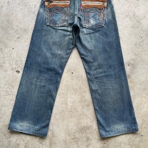May include: Blue denim jeans with brown leather accents on the back pockets. The jeans have a faded wash and a classic five-pocket design. The back pockets feature a unique stitched detail.