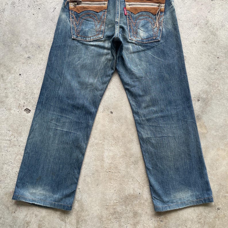 May include: Blue denim jeans with brown leather accents on the back pockets. The jeans have a faded wash and a classic five-pocket design. The back pockets feature a unique stitched detail.