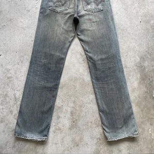 Puede incluir: Vaqueros de mezclilla gris descolorido con un dise&ntilde;o cl&aacute;sico de cinco bolsillos. Los vaqueros tienen un corte de pierna recta y presentan las costuras caracter&iacute;sticas de los bolsillos traseros. La mezclilla tiene una apariencia desgastada, lo que sugiere un art&iacute;culo vintage o de segunda mano.