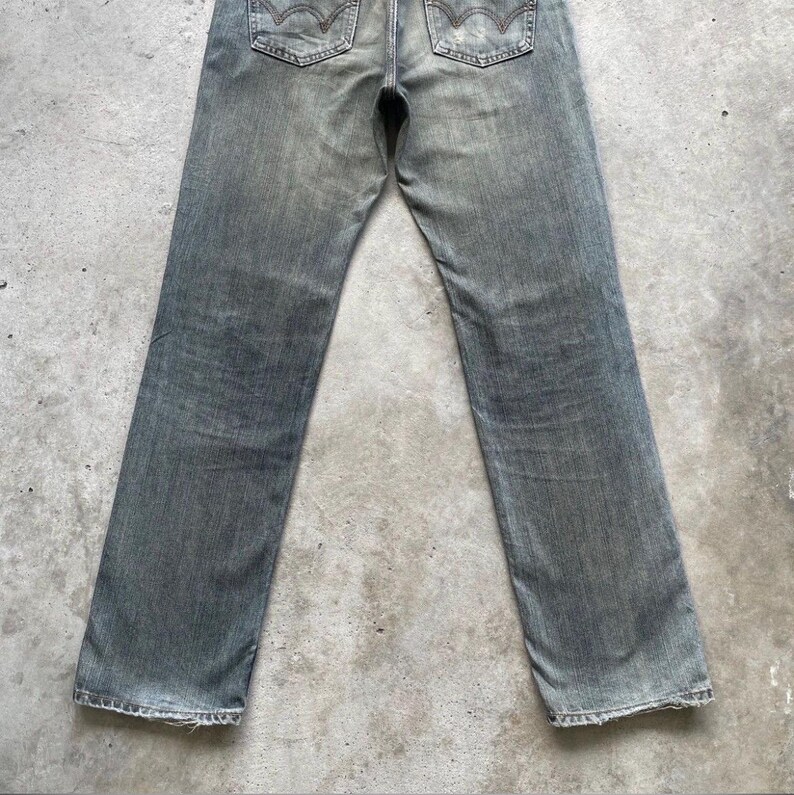 Puede incluir: Vaqueros de mezclilla gris descolorido con un dise&ntilde;o cl&aacute;sico de cinco bolsillos. Los vaqueros tienen un corte de pierna recta y presentan las costuras caracter&iacute;sticas de los bolsillos traseros. La mezclilla tiene una apariencia desgastada, lo que sugiere un art&iacute;culo vintage o de segunda mano.