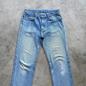 May include: Light blue denim jeans with a classic five-pocket design. The jeans have a faded wash and a straight leg cut. The image shows the jeans laid flat on a gray surface, showcasing the front of the garment.