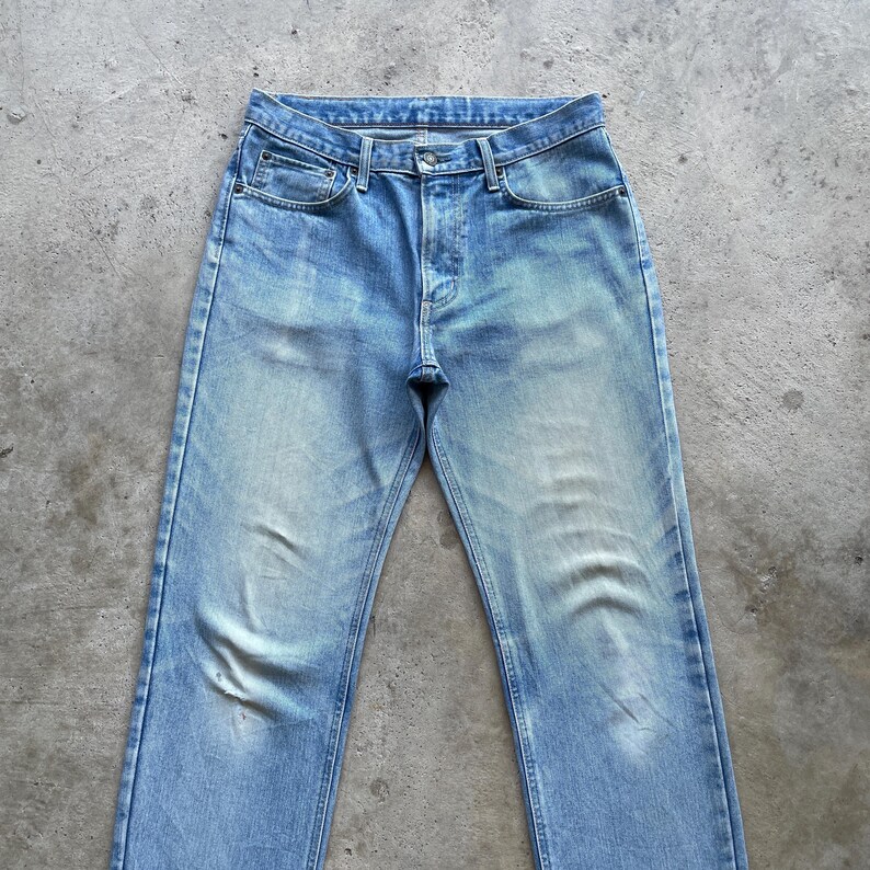 May include: Light blue denim jeans with a classic five-pocket design. The jeans have a faded wash and a straight leg cut. The image shows the jeans laid flat on a gray surface, showcasing the front of the garment.
