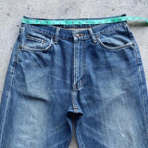 Pu&ograve; includere: Primo piano di jeans blu con un metro a nastro sulla vita. I jeans hanno un classico design a cinque tasche, una chiusura con bottoni e una cerniera. La vita misura circa 40,6 cm di larghezza.