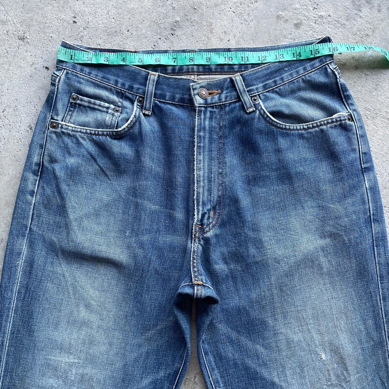 Pu&ograve; includere: Primo piano di jeans blu con un metro a nastro sulla vita. I jeans hanno un classico design a cinque tasche, una chiusura con bottoni e una cerniera. La vita misura circa 40,6 cm di larghezza.