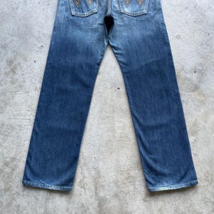 May include: Blue denim jeans with a faded wash. The jeans have a classic five-pocket design and feature decorative stitching on the back pockets. The jeans are laid flat on a concrete surface.
