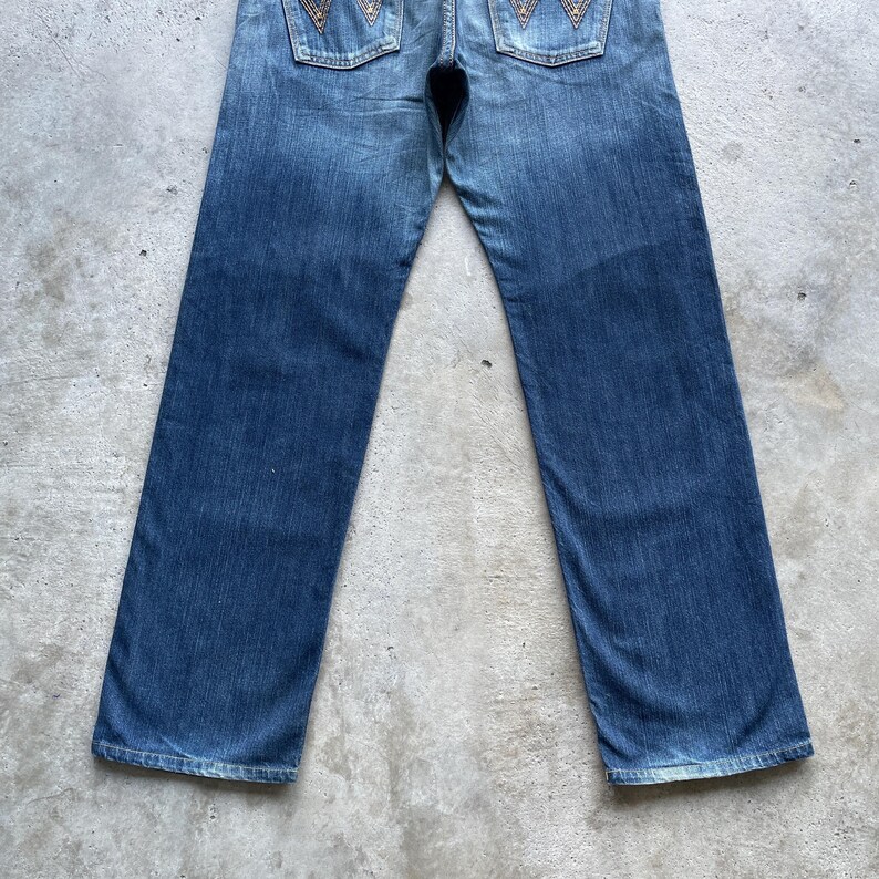 May include: Blue denim jeans with a faded wash. The jeans have a classic five-pocket design and feature decorative stitching on the back pockets. The jeans are laid flat on a concrete surface.