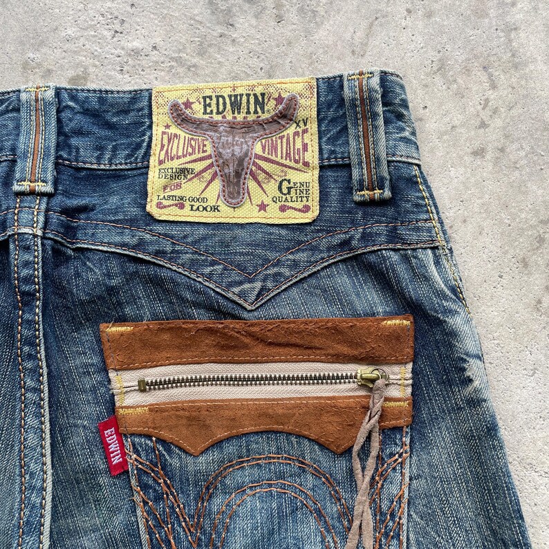May include: Close-up of blue denim jeans with a brown leather pocket featuring a zipper. The jeans have a vintage look with a faded wash and the brand name "EDWIN" is visible on a leather patch.