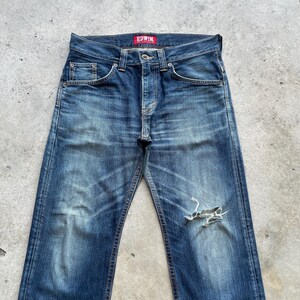 May include: Faded blue denim jeans with a distressed appearance, including a ripped knee. The jeans have a classic five-pocket design, a button and zip closure, and the brand name "EDWIN" on a red tag.