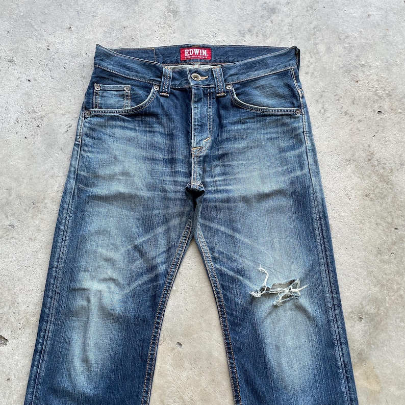 May include: Faded blue denim jeans with a distressed appearance, including a ripped knee. The jeans have a classic five-pocket design, a button and zip closure, and the brand name "EDWIN" on a red tag.
