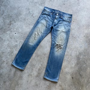 Puede incluir: Vaqueros de mezclilla azul descolorido con un aspecto desgastado, con una rodilla rasgada y el texto "503 Slim" en la cintura. Los vaqueros tienen un dise&ntilde;o cl&aacute;sico de cinco bolsillos y bragueta con botones.