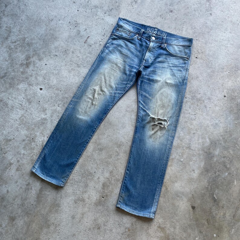 Puede incluir: Vaqueros de mezclilla azul descolorido con un aspecto desgastado, con una rodilla rasgada y el texto "503 Slim" en la cintura. Los vaqueros tienen un dise&ntilde;o cl&aacute;sico de cinco bolsillos y bragueta con botones.