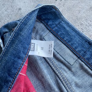Puede incluir: Primer plano de unos vaqueros de mezclilla con una etiqueta visible. La etiqueta muestra la marca "Edwin", junto con la talla "32" y otros c&oacute;digos de producto. Los vaqueros son de un azul medio con costuras visibles.