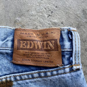 May include: Close-up of blue denim jeans with a brown leather patch. The patch reads "MADE IN U.S.A. LIMITED VERSION" and features the brand name "EDWIN". The text "THIS GARMENT HAS BEEN MADE IN USA UNDER STRICT QUALITY SUPERVISION EDWIN COMPANY LIMITED" is also visible.