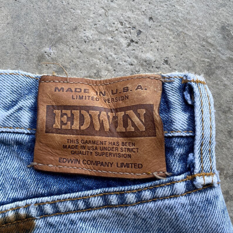 May include: Close-up of blue denim jeans with a brown leather patch. The patch reads "MADE IN U.S.A. LIMITED VERSION" and features the brand name "EDWIN". The text "THIS GARMENT HAS BEEN MADE IN USA UNDER STRICT QUALITY SUPERVISION EDWIN COMPANY LIMITED" is also visible.