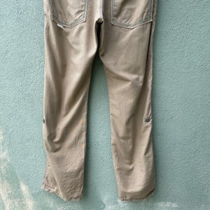 May include: Khaki trousers with visible stitching and two back pockets. The trousers have a straight leg design and a button detail near the knee. The stitching is a contrasting colour, adding a subtle design element to the garment.