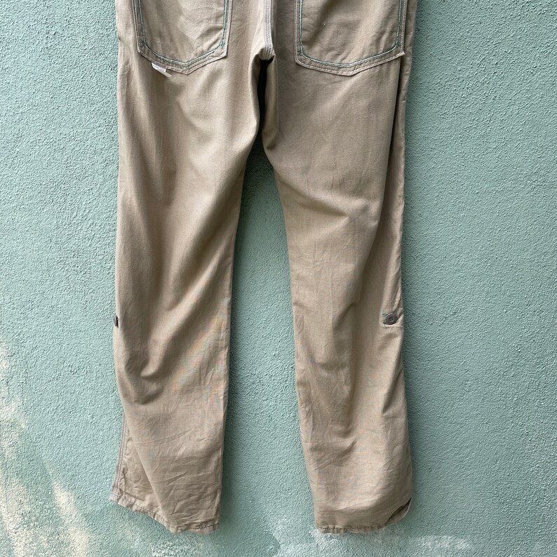 May include: Khaki trousers with visible stitching and two back pockets. The trousers have a straight leg design and a button detail near the knee. The stitching is a contrasting colour, adding a subtle design element to the garment.