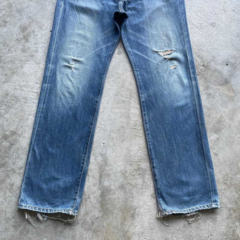 Pu&ograve; includere: Jeans in denim blu sbiadito con dettagli strappati. I jeans hanno un classico design a cinque tasche, una chiusura con bottone e cerniera e una vestibilit&agrave; dritta. Il denim mostra segni di usura, con alcune aree sbiadite e strappate.