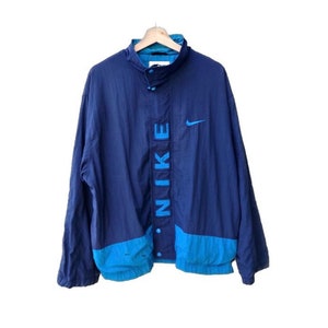 Puede incluir: Chaqueta cortavientos Nike azul marino vintage con un acento azul claro en la parte inferior y el cuello. La palabra "NIKE" se muestra verticalmente en la parte delantera, con el logo de Nike en el pecho derecho. La chaqueta está abotonada y colgada en una percha de madera.