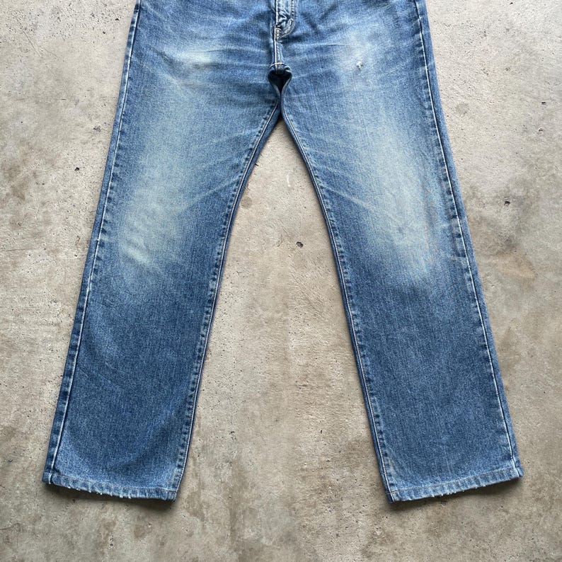May include: Faded blue denim jeans with a classic straight-leg fit. The jeans show signs of wear, with lighter patches and subtle distressing. The five-pocket design and button closure are visible.