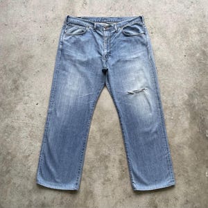 K&ouml;nnte beinhalten: Hellblaue Jeans mit einem abgenutzten, gebrauchten Aussehen. Die Jeans haben ein klassisches F&uuml;nf-Taschen-Design, einen Knopf- und Rei&szlig;verschluss und einen kleinen Riss am rechten Bein. Die Jeans liegen flach auf einer Betonoberfl&auml;che.