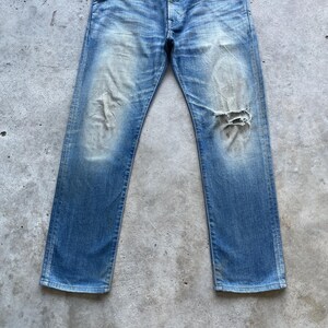 Puede incluir: Vaqueros de mezclilla azul descolorido con un aspecto desgastado. Los vaqueros tienen un aspecto usado con un gran desgarro en la rodilla derecha. La mezclilla tiene variaciones de azul claro y oscuro, creando una est&eacute;tica vintage.