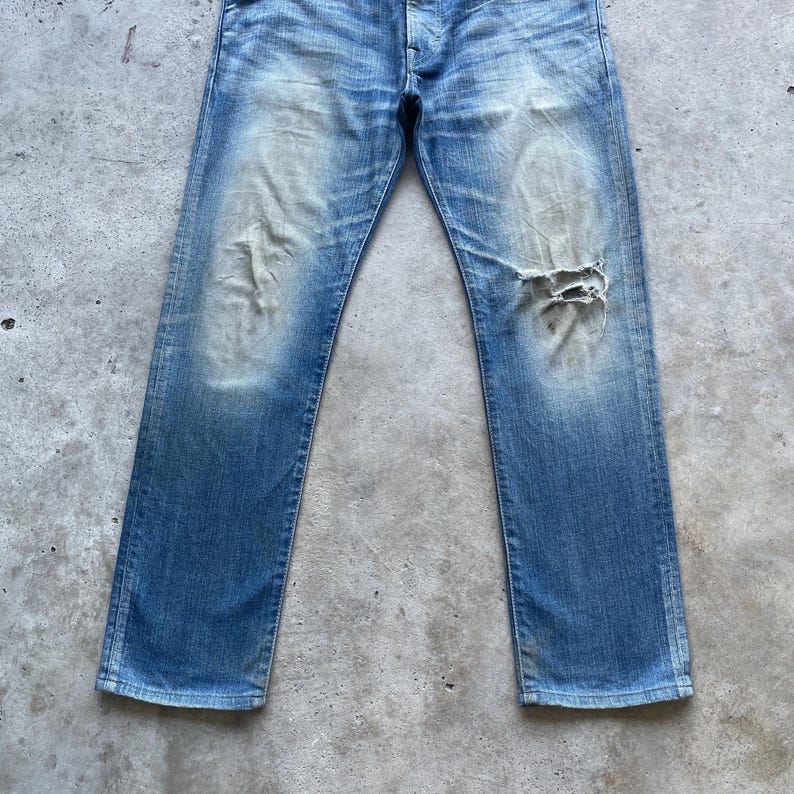 Puede incluir: Vaqueros de mezclilla azul descolorido con un aspecto desgastado. Los vaqueros tienen un aspecto usado con un gran desgarro en la rodilla derecha. La mezclilla tiene variaciones de azul claro y oscuro, creando una est&eacute;tica vintage.